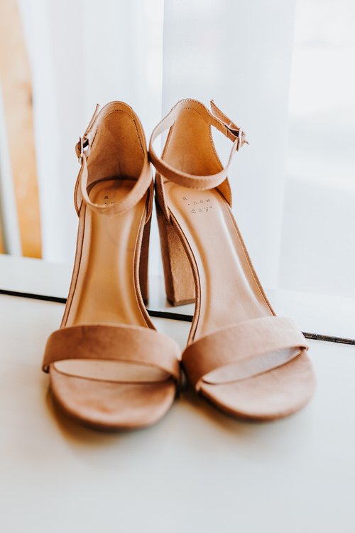 Pair of light brown suede block-heeled sandals with ankle straps on a white surface, with a window in the background.