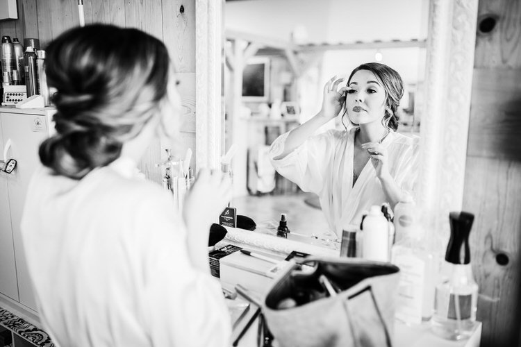 Bride in a white robe applies makeup, looking into a mirror in a rustic cabin interior. Reflections show wooden beams.