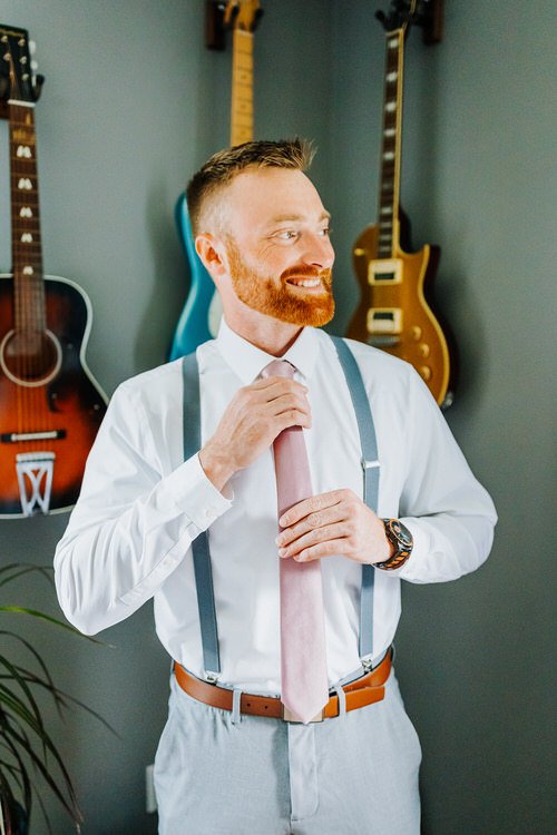 Man in white shirt, pink tie, grey suspenders adjusts his tie. Three guitars hang on a grey wall behind him.