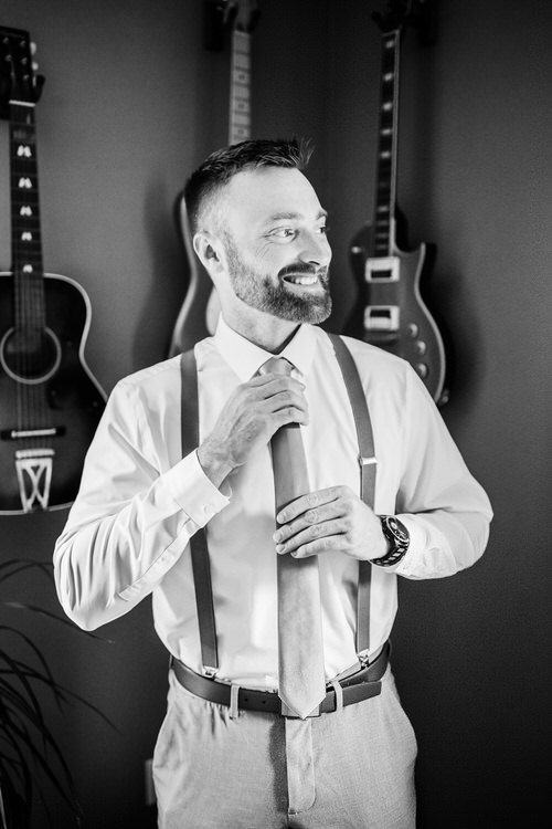 Black and white photo of a bearded man adjusting his tie, wearing a white shirt and suspenders, with three guitars on the...