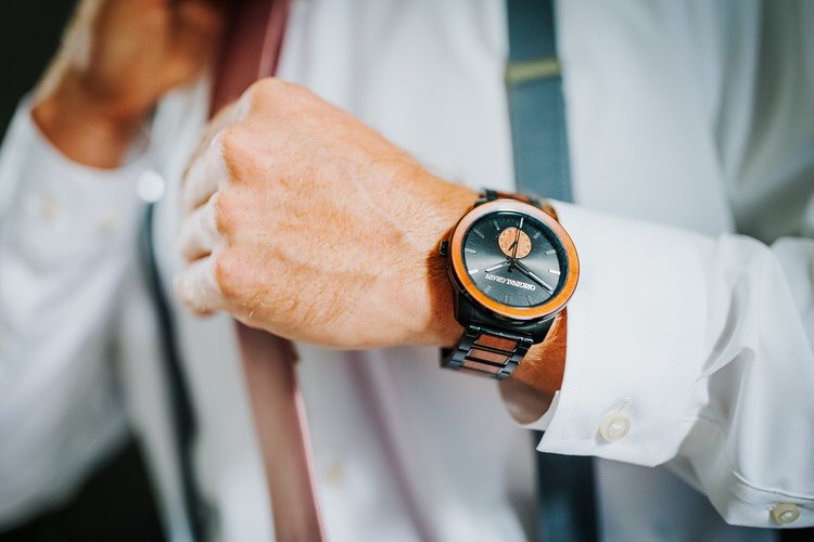 A person's hand adjusts a tie while wearing a watch with a wooden bezel and band, and a white shirt with French cuffs.