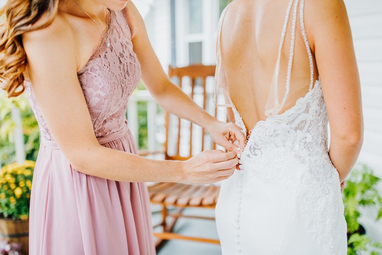 Person in pink dress buttons up the back of a white wedding gown with pearl straps. Outdoor porch setting with rocking chair.