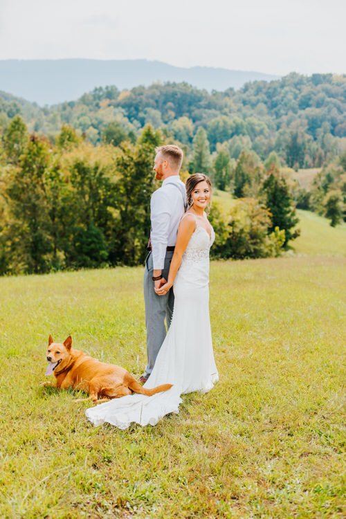 Bride, groom, and a brown dog pose in a grassy field with green mountains and trees in the background. They stand...