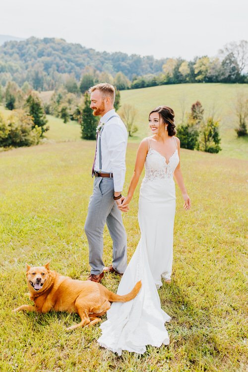 A couple holding hands in a grassy field with their dog, against a backdrop of rolling hills and trees at Moose Hollow Lodge.