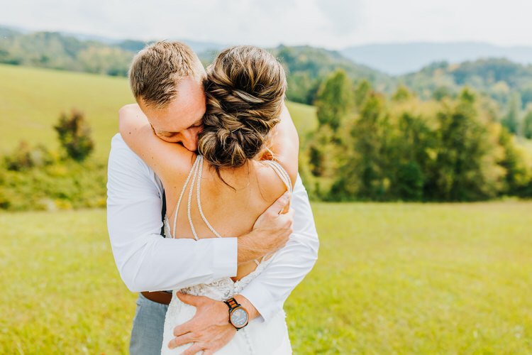 Man in white shirt embraces woman in white wedding dress with pearl straps in a grassy field with mountains.