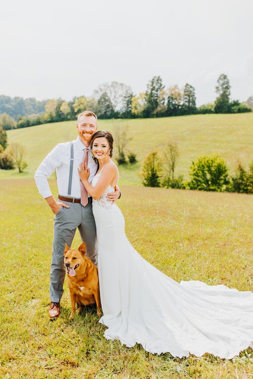 Wedding couple and a dog in a grassy field with rolling hills and trees in the background under a light sky.