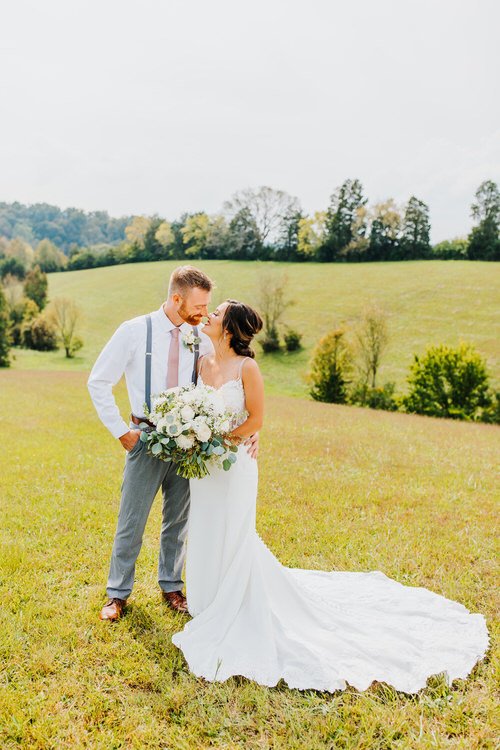 Bride in white dress with train and groom in white shirt, suspenders, pink tie stand in grassy field with rolling hills...