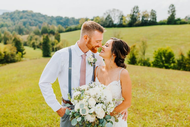 Wedding couple smiles in a grassy field with rolling hills and trees. Groom wears suspenders, bride holds a white bouquet.