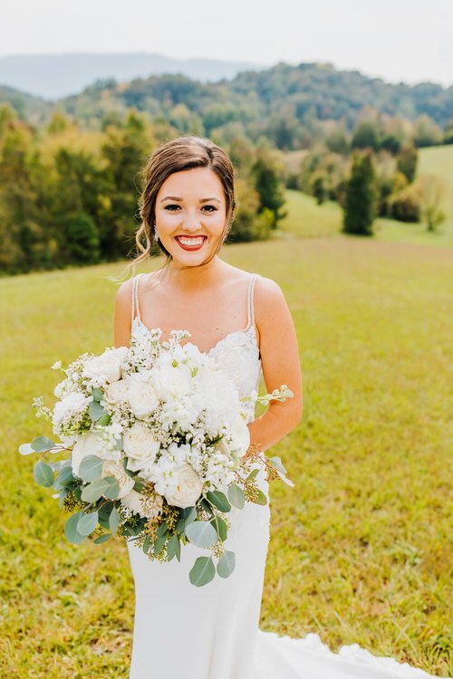 Smiling bride in a white wedding dress holds a large white and green bouquet in a grassy field at Moose Hollow Lodge with...