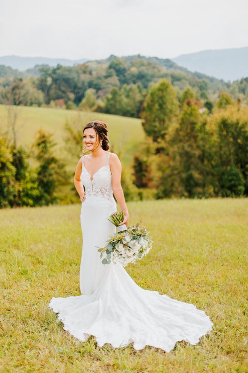 Bride in a white gown holding a bouquet stands in a grassy field with rolling hills and trees in the background.