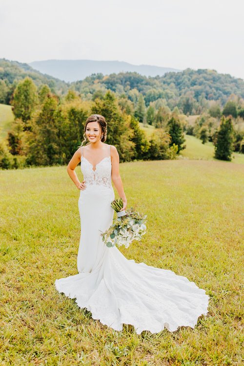 Bride in white dress holding a bouquet in a grassy field with rolling hills and trees in the background under natural light.