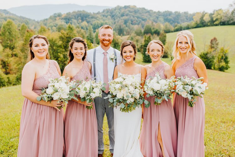 A wedding party, including a bride, groom, and four bridesmaids, stands outdoors in a grassy field with rolling hills and...