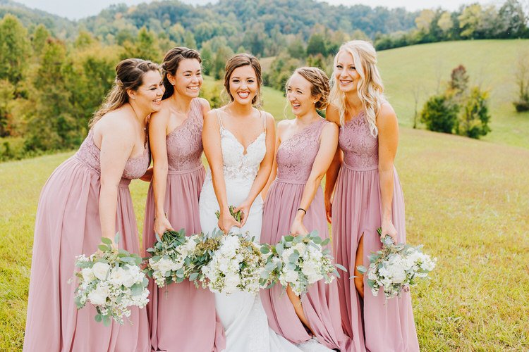 Bride in white gown and four bridesmaids in dusty rose dresses hold bouquets in a grassy field with green hills.