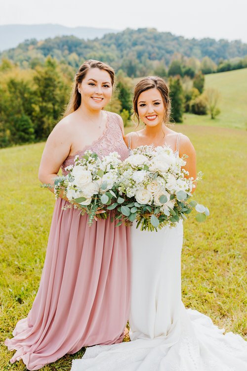 Bride in white dress and bridesmaid in dusty rose dress holding bouquets in a grassy field with green hills background.