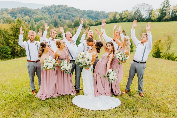 Wedding couple kissing surrounded by 10 members of wedding party in a field with hills and trees. Bridesmaids in pink,...