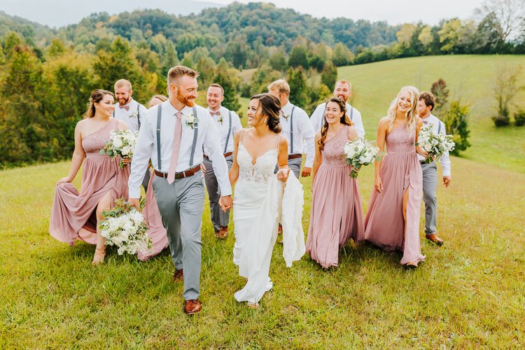Wedding party walks in grassy field with bride, groom, bridesmaids in mauve dresses, groomsmen in suspenders. Mountain...