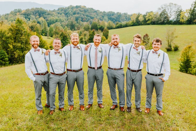 Seven groomsmen in white shirts, grey pants, suspenders, and bow ties stand on a grassy hill with a forested mountain...