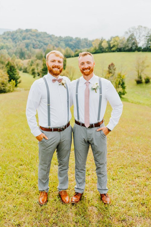 Two men in white shirts, grey pants, suspenders, and brown shoes stand in a grassy field with hills.