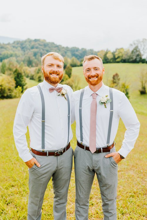Two men in white shirts, grey suspenders, grey pants, and pink accessories stand in a grassy field with green hills.