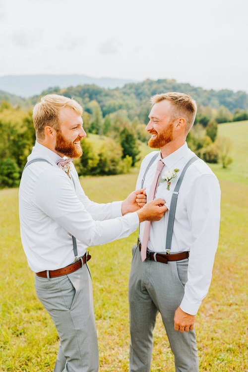 Two men in white shirts and grey pants outdoors, one adjusting the other's boutonnière. Green hills and distant mountains...