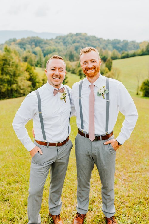 Two men in white shirts, grey pants, suspenders, and boutonnieres stand smiling in a grassy field with rolling hills.