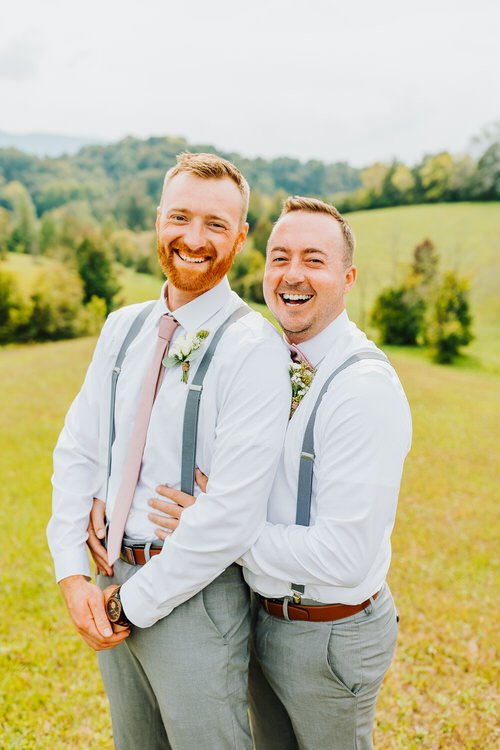 Two men in white shirts, pink ties, and grey suspenders smile outdoors at Moose Hollow Lodge with hills behind them.
