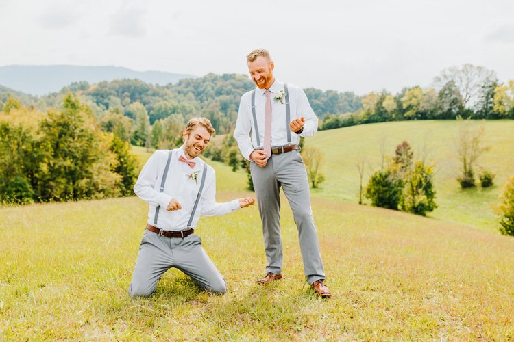 Two men in white shirts, gray pants, and suspenders pose with air guitars in a grassy field with rolling hills.