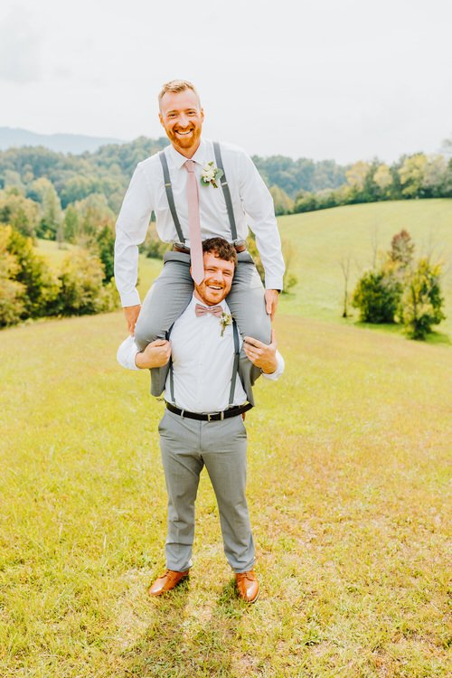 Two men in wedding attire, one on the other's shoulders, smiling in a grassy field with green hills at Moose Hollow Lodge.