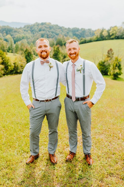 Two men in white shirts, gray pants, suspenders, and boutonnieres stand in a grassy field with green hills behind them.