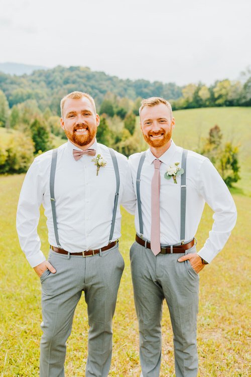 Two men in white shirts, grey pants, and pink accessories stand outdoors at Moose Hollow Lodge with green hills.