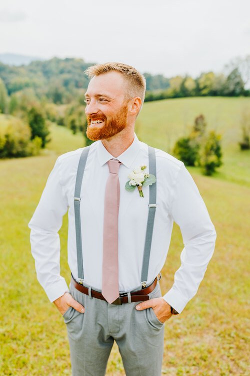 Groom in white shirt, grey suspenders, dusty rose tie, and boutonniere, smiling outdoors in a green field with hills.