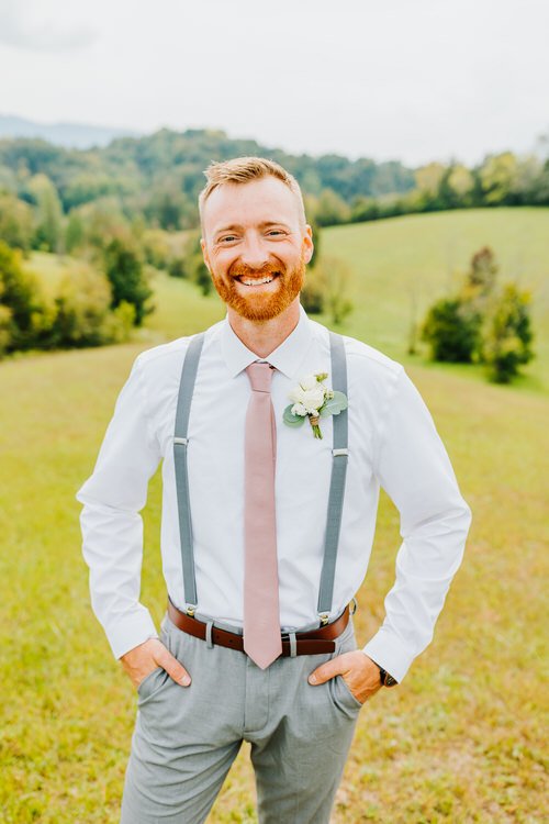 Man in white shirt, pink tie, grey suspenders, and grey pants with a boutonniere, smiling. Outdoors with green hills and...