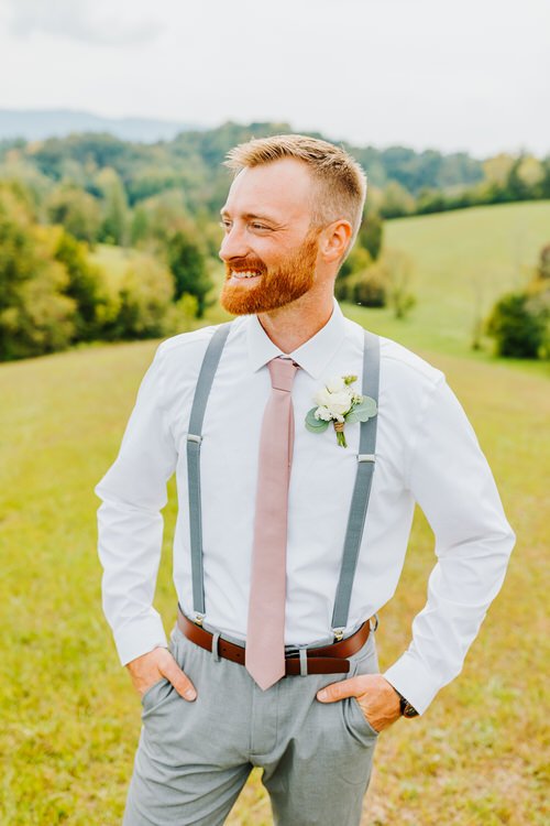 Man in white shirt, pink tie, and gray suspenders and pants, standing outdoors with green rolling hills and mountains...