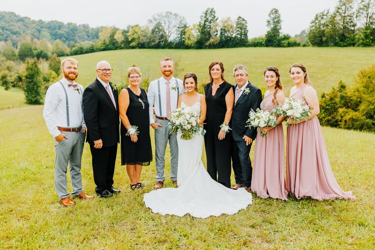 Wedding party and family stand in a grassy field with rolling green hills and trees in the background.