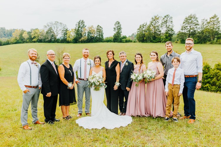 Wedding party and family stand in a grassy field with trees and hills in the background under an overcast sky.