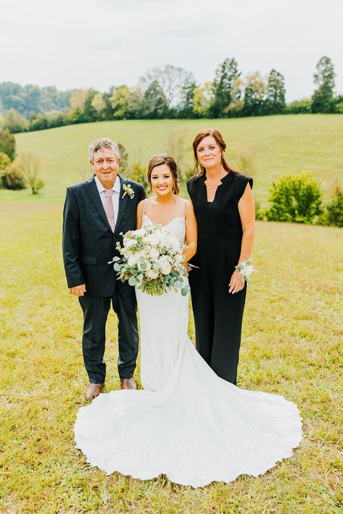 Bride in a white wedding dress with parents in a grassy field with trees and hills in the background.