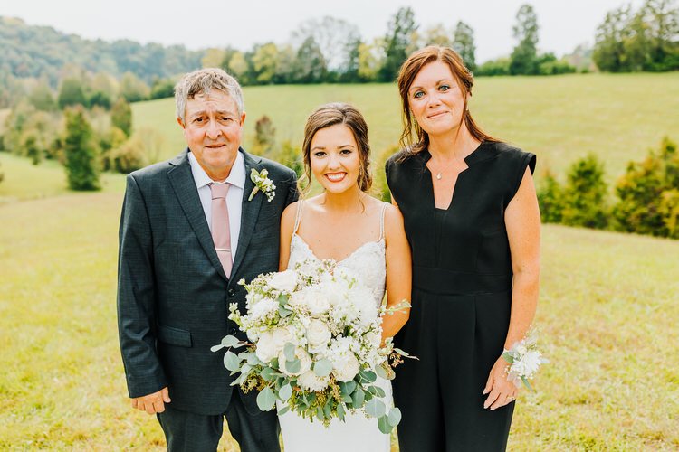 Bride in white dress holds large white bouquet, flanked by man in suit and woman in black. Outdoors in green field with...