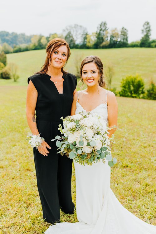 Two women, one in a white wedding dress with a white bouquet and one in a black jumpsuit, stand in a grassy field with...