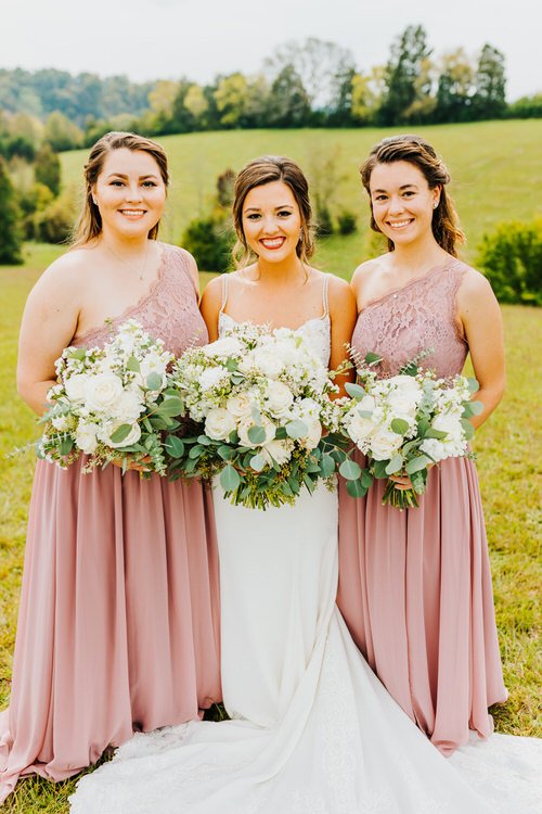 Bride in white dress with two bridesmaids in dusty rose dresses hold bouquets in a grassy field with trees in background.