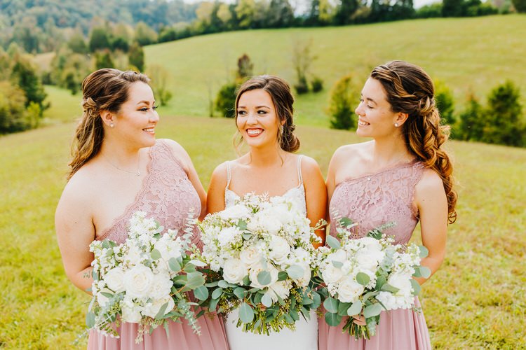 Bride in white dress and two bridesmaids in pink dresses with bouquets in a grassy field at Moose Hollow Lodge.