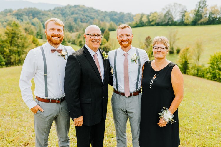 Four adults, dressed in wedding attire, stand in a sunny green field with distant tree-covered mountains.