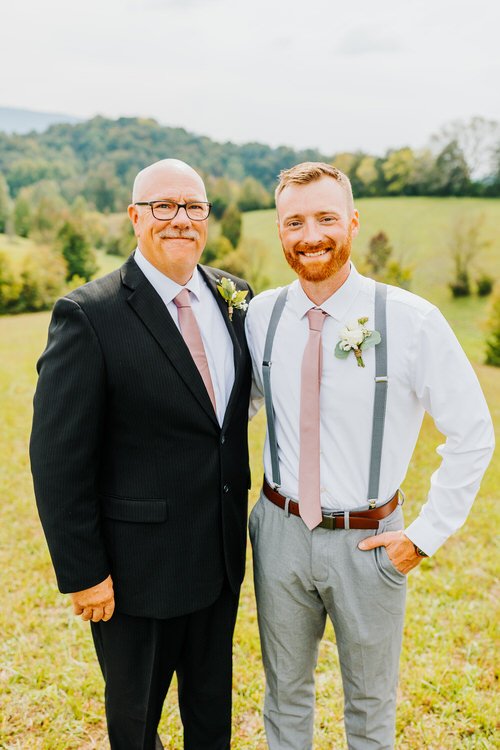 Two men in wedding attire pose outdoors in a grassy field with distant green hills and trees under a bright sky.