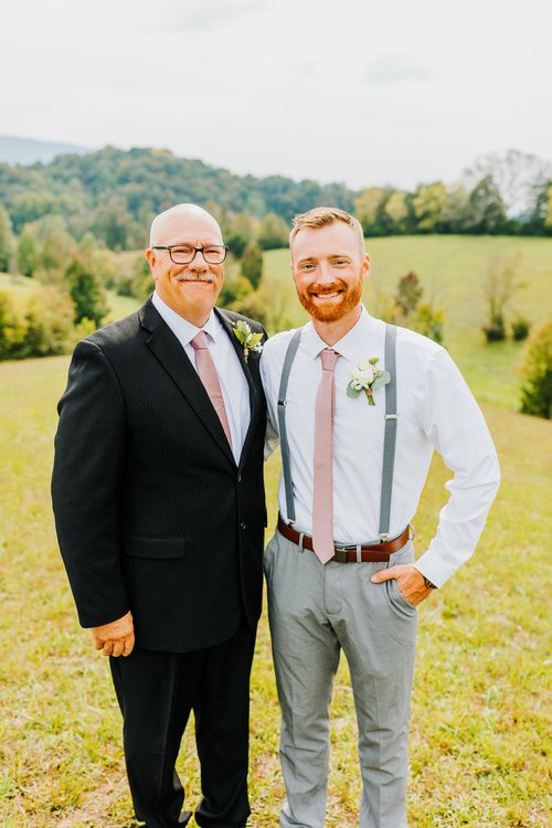 Two men in formal attire stand in a grassy field with rolling hills and trees in the background. Moose Hollow Lodge setting.