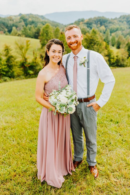 Couple in formal wear stand on grassy hill, woman holding white and green bouquet. Green wooded hills in background.
