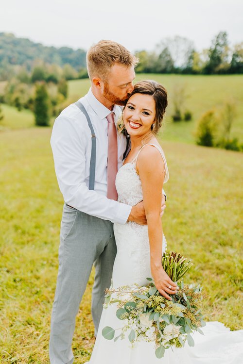 Couple in wedding attire posing in a grassy field with rolling hills in the background. Groom kisses bride's forehead.