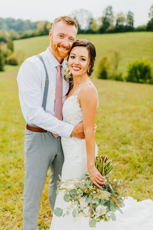 Bride and groom embrace in a grassy field, smiling at the camera. Groom wears gray pants, white shirt, suspenders; bride...