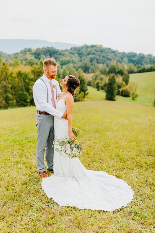 Bride and groom embrace in a grassy field with rolling green hills and trees in the background under a light sky.