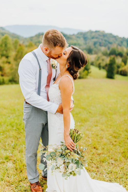 Couple kissing in a grassy field. Woman in white dress holds a bouquet. Man in white shirt, gray suspenders, pink tie....
