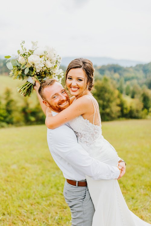 Groom lifting bride in a grassy field with a white and green bouquet. Hills and trees are in the background under an...
