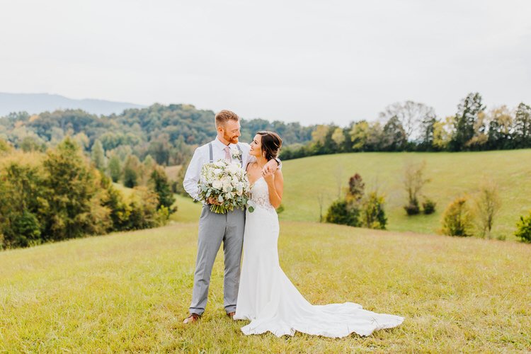 Couple stands in a grassy field with rolling hills and trees in the background. Groom holds a large white bouquet.